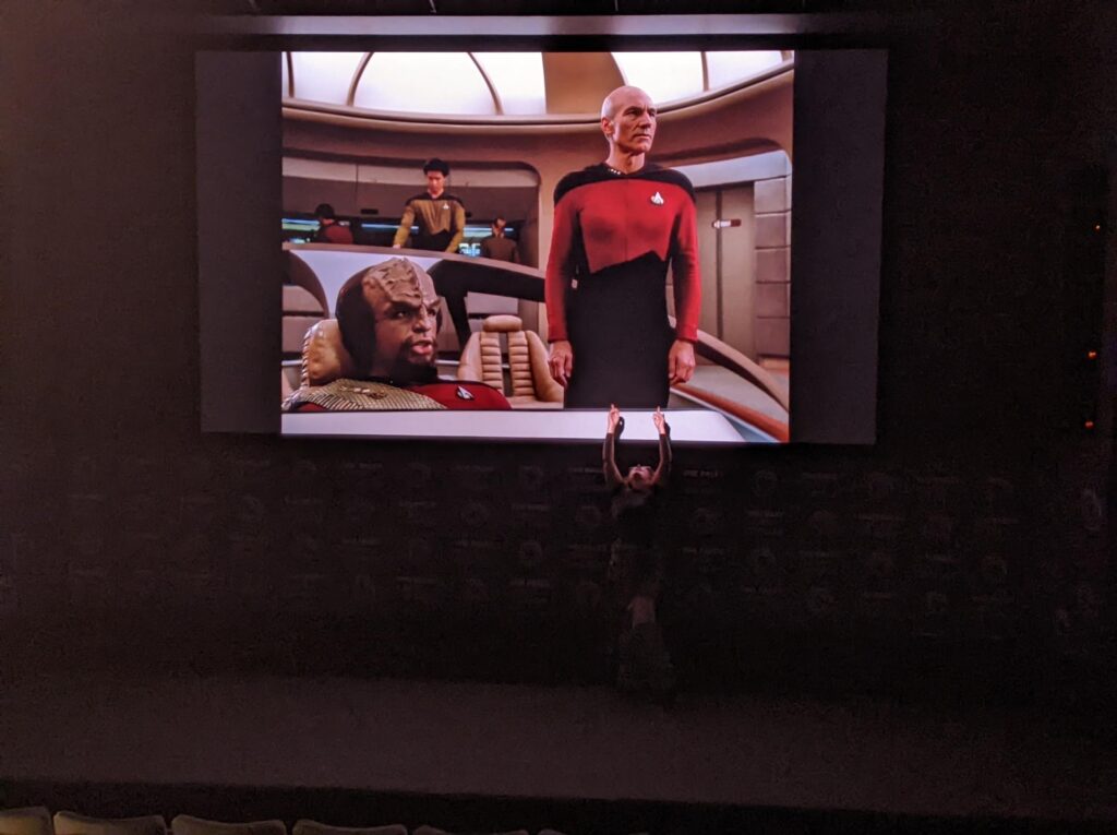 Sharon Simon on stage while an episode of Star Trek TNG plays in the background. She's pointing at Captain Picard.