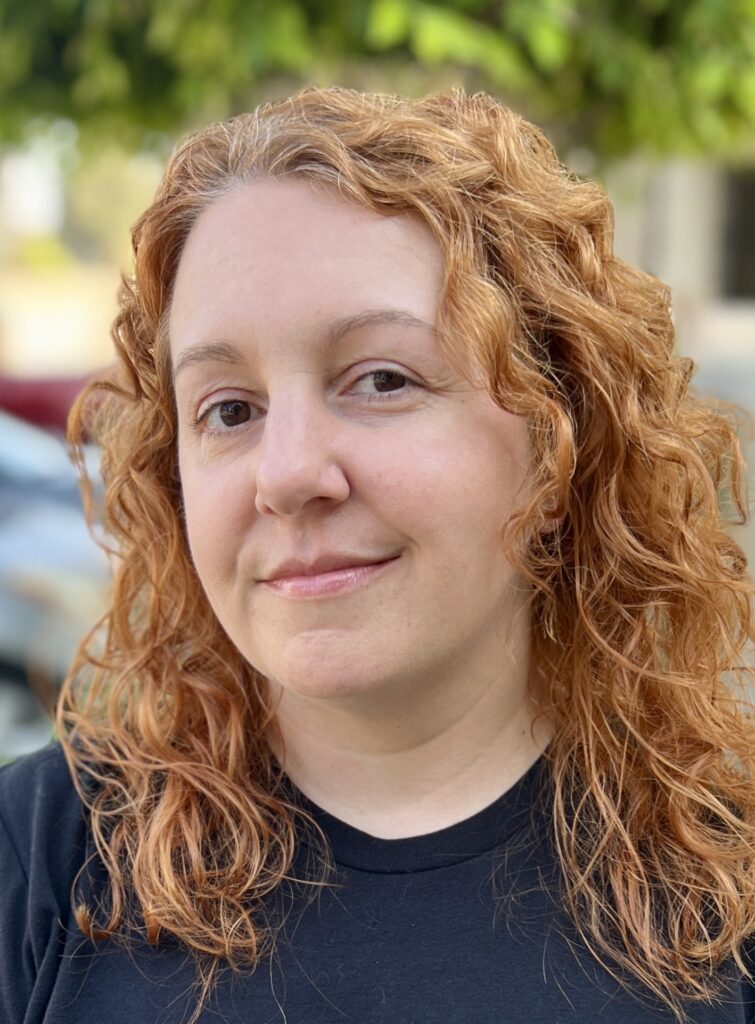 headshot of Charlynn Schmiedt, a red-headed woman wearing a dark tee shirt with a nature background