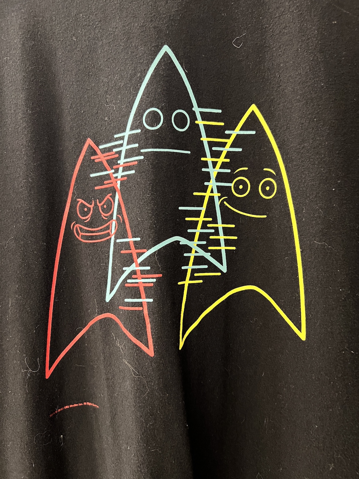 A black tee shirt with three Badgeys from Star Trek Lower Decks. There is one in the middle with a neutral expression and then a red angry badgey on the left and a happy yellow badget on the right.