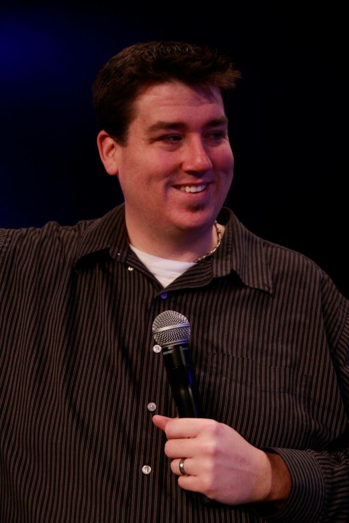 Image of Brent Allen wearing a brown and black striped shirt. He is smiling and holding a microphone.